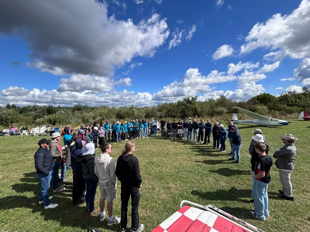 Teilnehmerinnen und Teilnehmer des Jugendvergleichsfliegens stehen im Kreis auf der Graspiste in Breitscheid, im Hintergrund ein Segelflugzeug