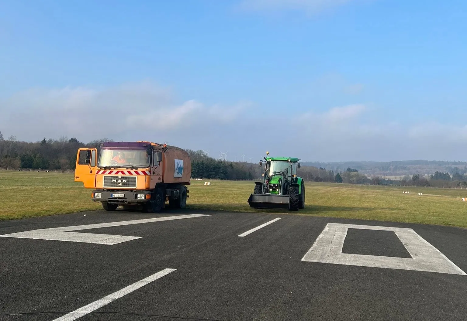 Orangefarbener MAN-Kehrwagen und grüner Traktor mit Frontlader am Rand der Piste unter freiem Himmel