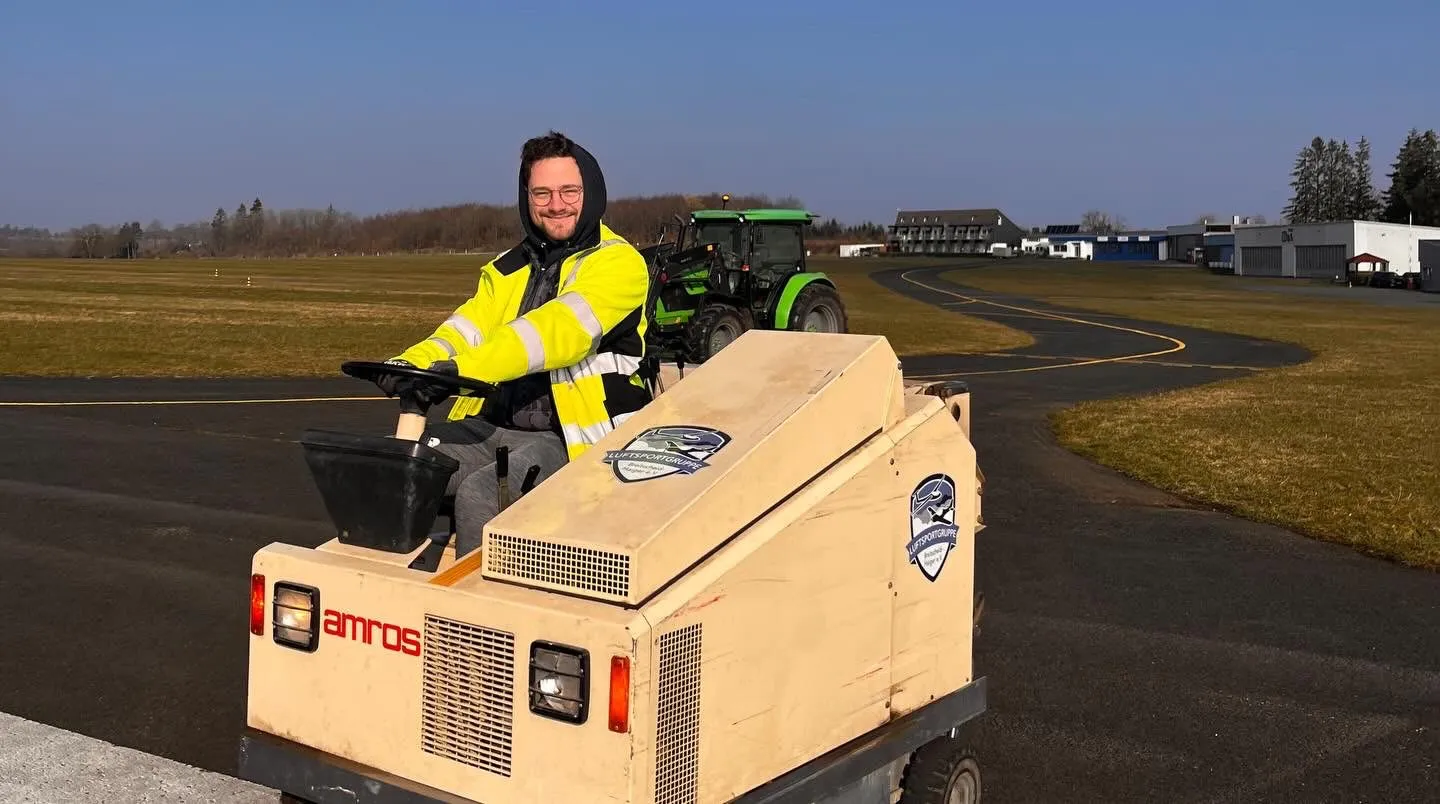 Vereinsmitglied bedient eine kompakte Kehrmaschine auf dem Rollweg, im Hintergrund Traktor und Flugplatzgebäude
